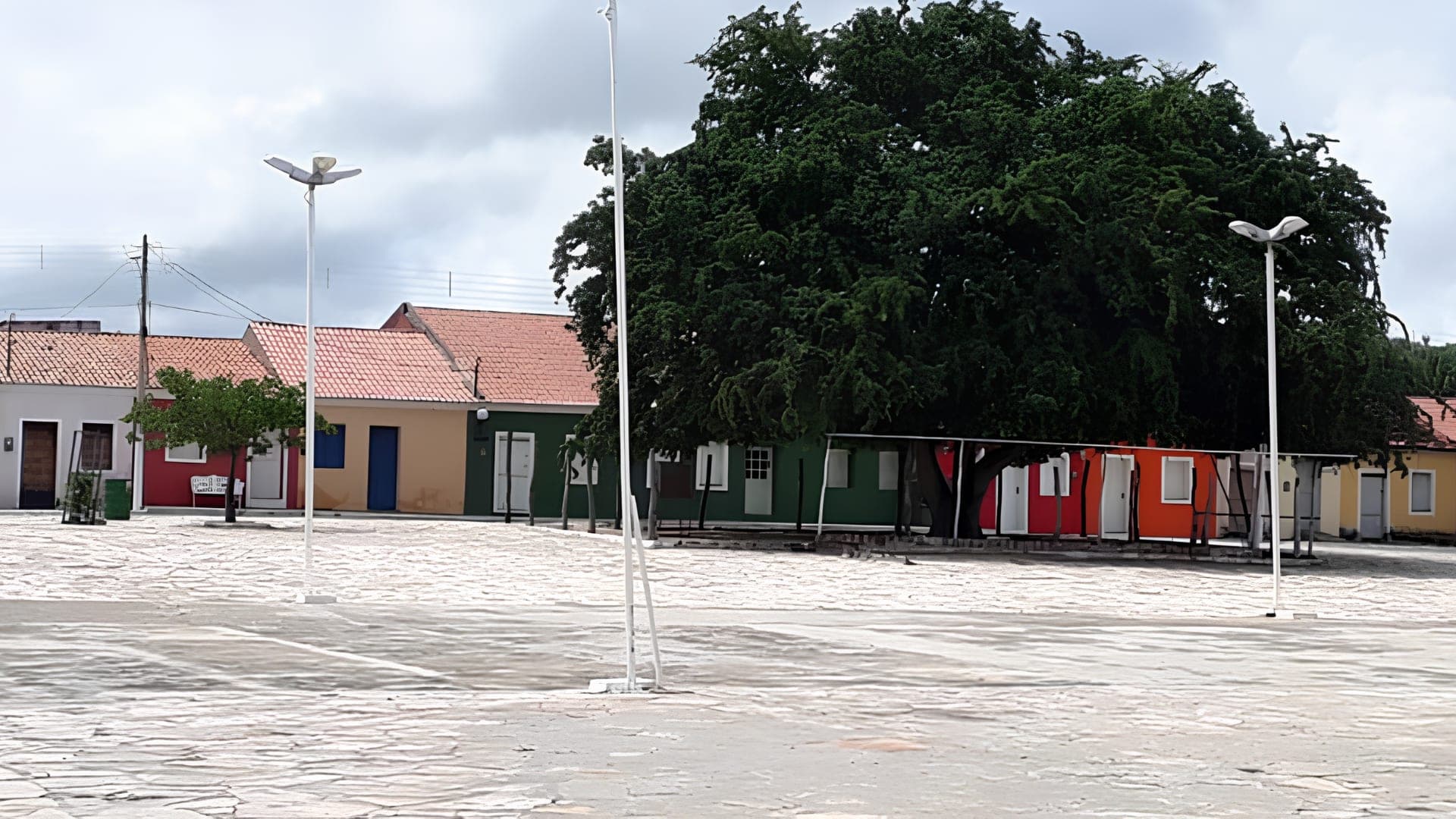 Largo do povoado de Caboclo, com piso de pedra, postes de iluminação e os casarios coloridos ao fundo sob um tamarineiro.