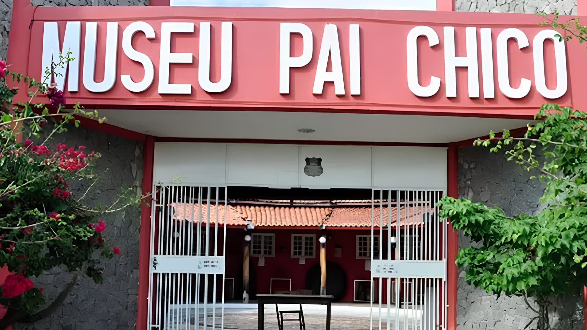 Fachada do Museu Pai Chico, com letreiro em relevo, portão de grades brancas e primaveras floridas nas laterais.